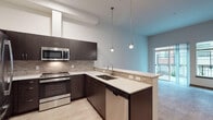 This single-floor residence showcases a thoughtfully designed layout with a cohesive modern aesthetic. The kitchen serves as a standout feature, equipped with a full suite of stainless steel GE appliances, including a French door refrigerator, electric range, built-in microwave, and dishwasher. Rich dark-toned cabinetry contrasts elegantly with light quartz countertops, while a textured tile backsplash adds visual depth. A peninsula with an undermount double sink opens toward the living area, illuminated by stylish pendant lighting suspended from above.

The living area is bright and open, with light wood-look flooring, tall ceilings, and sliding glass doors leading to a private outdoor balcony. Natural light flows generously through the large glazed door and adjacent window, creating an airy atmosphere.

The bedroom features plush carpeted flooring and two large windows fitted with horizontal blinds, offering views of the surrounding exterior and allowing ample daylight. The bathroom maintains the same refined palette, with dark cabinetry, a wide undermount vanity sink, a large mirror, light wood-look tile flooring, and conveniently placed grab bars, suggesting accessibility-conscious design. Overall, the space presents a clean, contemporary feel throughout.