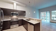 This single-floor residence showcases a thoughtfully designed layout with a cohesive modern aesthetic. The kitchen serves as a standout feature, equipped with a full suite of stainless steel GE appliances, including a French door refrigerator, electric range, built-in microwave, and dishwasher. Rich dark-toned cabinetry contrasts elegantly with light quartz countertops, while a textured tile backsplash adds visual depth. A peninsula with an undermount double sink opens toward the living area, illuminated by stylish pendant lighting suspended from above.

The living area is bright and open, with light wood-look flooring, tall ceilings, and sliding glass doors leading to a private outdoor balcony. Natural light flows generously through the large glazed door and adjacent window, creating an airy atmosphere.

The bedroom features plush carpeted flooring and two large windows fitted with horizontal blinds, offering views of the surrounding exterior and allowing ample daylight. The bathroom maintains the same refined palette, with dark cabinetry, a wide undermount vanity sink, a large mirror, light wood-look tile flooring, and conveniently placed grab bars, suggesting accessibility-conscious design. Overall, the space presents a clean, contemporary feel throughout.