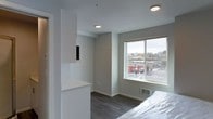 This single-floor space showcases a contemporary design with a neutral color palette of soft grays and crisp whites throughout. The living area features dark hardwood-style flooring that contrasts cleanly against the light-toned walls and white ceiling, creating a modern and cohesive aesthetic. Recessed lighting is positioned throughout the ceiling, providing consistent, even illumination across the space.

A large sliding window serves as a focal point, offering elevated views of the surrounding urban streetscape, including nearby commercial buildings and street-level activity. The natural light entering through this window brightens the interior considerably, enhancing the open feel of the space.

A built-in white partition wall adds architectural interest while also functioning as a subtle room divider, incorporating a pass-through cutout and a low countertop surface — useful for display or everyday tasks. A floor-mounted HVAC vent beneath the window ensures climate control. The overall ambiance is clean, modern, and understated, reflecting a practical yet well-finished design suited for urban living.