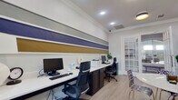 This single-floor business center is thoughtfully designed to support both productivity and collaboration. The workspace is anchored by an expansive built-in desk running along one wall, offering multiple computer workstations equipped with monitors, keyboards, and a shared printer. Dark cabinetry beneath the white countertop provides ample storage, while ergonomic task chairs ensure comfort during extended use. A striking horizontal striped accent wall in navy blue, silver-gray, and gold adds a bold, contemporary visual element that defines the character of the room. Crown molding and wainscoting lend a refined architectural finish throughout. Adjacent to the workstation area, a collaborative lounge zone features a round marble-top table surrounded by an eclectic mix of acrylic and patterned upholstered chairs, ideal for informal meetings or group discussions. Bold abstract artwork in a blue-and-white palette decorates the walls, reinforcing the space's modern aesthetic. Light wood-tone flooring flows continuously throughout, contributing to a bright and open feel. French doors with glass panels allow natural light to filter in from outside, while interior glass windows maintain visual connectivity between zones. Recessed lighting overhead ensures the space remains well-lit at all hours.