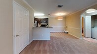 This single-story residence features a well-organized layout that connects functional living spaces in a cohesive design. The galley-style kitchen is outfitted with dark espresso cabinetry, granite-look countertops, and black appliances including a refrigerator, electric range, and microwave. Wood-look plank flooring runs underfoot, adding warmth and easy maintenance to the cooking space. A peninsula counter extends into the living area, creating an open-concept feel that enhances the sense of flow between the kitchen and main living space. The living area is finished with plush neutral carpeting, crown molding, and warm beige walls, lending a comfortable, welcoming atmosphere. A pendant light fixture adds a decorative touch overhead. The bedroom is a well-proportioned, carpeted room featuring a ceiling fan with light kit and a window with blinds that allows natural light to filter in. The bathroom is finished with wood-look flooring, a dark vanity with granite-look countertops, a large mirror, multi-bulb vanity lighting, and a full tub-shower combination with tile surround. Throughout the unit, warm neutral tones, consistent finishes, and practical design choices create a cohesive and comfortable living environment.
