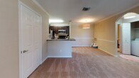 This single-story residence features a well-organized layout that connects functional living spaces in a cohesive design. The galley-style kitchen is outfitted with dark espresso cabinetry, granite-look countertops, and black appliances including a refrigerator, electric range, and microwave. Wood-look plank flooring runs underfoot, adding warmth and easy maintenance to the cooking space. A peninsula counter extends into the living area, creating an open-concept feel that enhances the sense of flow between the kitchen and main living space. The living area is finished with plush neutral carpeting, crown molding, and warm beige walls, lending a comfortable, welcoming atmosphere. A pendant light fixture adds a decorative touch overhead. The bedroom is a well-proportioned, carpeted room featuring a ceiling fan with light kit and a window with blinds that allows natural light to filter in. The bathroom is finished with wood-look flooring, a dark vanity with granite-look countertops, a large mirror, multi-bulb vanity lighting, and a full tub-shower combination with tile surround. Throughout the unit, warm neutral tones, consistent finishes, and practical design choices create a cohesive and comfortable living environment.