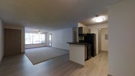 This single-floor space showcases an open-concept design that integrates the kitchen and living area into one cohesive layout. The kitchen is equipped with dark cabinetry, black appliances including a refrigerator and range, and a peninsula with a dark countertop that serves as a natural divider between the cooking and living zones. The kitchen flooring is light hardwood-style, which transitions into carpeting in the living area, adding warmth underfoot. The living space is generously sized, anchored by a ceiling fan and illuminated by a large front-facing window that allows ample natural light to fill the room. The textured popcorn ceiling is consistent throughout, and flush-mount lighting provides additional illumination. A hallway visible near the kitchen leads to additional areas of the unit, including what appears to be a closet with louvered doors. The neutral white walls throughout create a clean, versatile backdrop. Overall, the space offers a functional and straightforward layout with a clear separation of living and cooking zones defined by flooring materials and the kitchen peninsula.