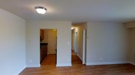 This single-story space features a straightforward, functional layout designed for practical everyday living. The main living area is finished with warm hardwood flooring that extends throughout, providing a cohesive look across the space. Neutral beige walls and white baseboards create a clean, understated backdrop throughout.

A dedicated kitchen is accessible through a doorway from the main area and is equipped with wooden cabinetry, dark countertops, and black appliances, including a refrigerator and stove. The kitchen benefits from its own overhead lighting and offers ample counter space for food preparation.

From the main area, a hallway extends toward the back of the unit, lined with multiple doors that lead to additional rooms. The hallway is well-lit with a ceiling-mounted light fixture, maintaining a bright and accessible feel. A bi-fold door, likely concealing a closet, is positioned at the entrance to the hallway, offering convenient storage. Flush-mount ceiling lighting is consistent throughout, ensuring even illumination across the space. The overall ambiance is clean, neutral, and move-in ready.