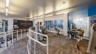 Located at 2101 Market St in Denver's vibrant RiNo neighborhood, this fitness center offers a thoughtfully designed, single-floor layout that accommodates a comprehensive range of workout options. The space is divided into distinct functional zones, each tailored to a specific training style, and connected by a sleek railing system that guides members through the facility.

The cardio zone features a lineup of treadmills, ellipticals, and stationary bikes positioned near large windows, allowing natural light to fill the area and providing views of the surrounding streetscape. The strength training section is generously stocked with Precor equipment, multi-rack cable machines, and a full array of dumbbells neatly arranged on tiered racks, all set atop durable hardwood-style flooring with protective rubber matting in key lifting areas.

A dedicated functional training zone features an open floor plan with carpet tile flooring, ceiling fans, and motivational chalkboard wall accents that add a personalized, energetic touch to the space. Overhead, linear fluorescent lighting ensures the entire facility is brightly and evenly lit throughout. Large interior windows and mirrors strategically placed along the walls enhance the sense of openness and allow for form-checking during workouts. The overall ambiance is clean, modern, and purposeful, creating an inviting environment for all fitness levels.