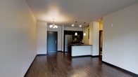 This single-floor space showcases a thoughtfully designed open-concept layout that integrates the living, dining, and kitchen areas into one cohesive environment. Dark hardwood flooring runs throughout, lending a warm and sophisticated tone to the interior. A decorative chandelier anchors the dining area, complementing the recessed lighting and sleek pendant lights that illuminate the kitchen space.

The kitchen is a standout feature, outfitted with dark upper cabinetry, dark countertops, and stainless steel appliances including a refrigerator and range with overhead microwave. A peninsula with a white base provides a casual breakfast bar or prep area, adding both functionality and visual contrast to the darker upper elements. A single-basin sink sits along the peninsula, positioned beneath pendant lighting for practical task illumination.

The front entry, framed by a dark-paneled door, transitions smoothly into the open living area, maintaining a clean sightline across the space. Neutral-toned walls keep the interior bright and versatile. A hallway visible toward the rear of the unit suggests additional rooms beyond the main living area, offering a sense of depth and privacy within this well-appointed single-level space.