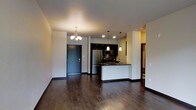 This single-floor space showcases a thoughtfully designed open-concept layout that integrates the living, dining, and kitchen areas into one cohesive environment. Dark hardwood flooring runs throughout, lending a warm and sophisticated tone to the interior. A decorative chandelier anchors the dining area, complementing the recessed lighting and sleek pendant lights that illuminate the kitchen space.

The kitchen is a standout feature, outfitted with dark upper cabinetry, dark countertops, and stainless steel appliances including a refrigerator and range with overhead microwave. A peninsula with a white base provides a casual breakfast bar or prep area, adding both functionality and visual contrast to the darker upper elements. A single-basin sink sits along the peninsula, positioned beneath pendant lighting for practical task illumination.

The front entry, framed by a dark-paneled door, transitions smoothly into the open living area, maintaining a clean sightline across the space. Neutral-toned walls keep the interior bright and versatile. A hallway visible toward the rear of the unit suggests additional rooms beyond the main living area, offering a sense of depth and privacy within this well-appointed single-level space.