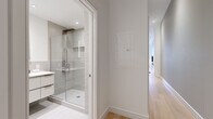 This thoughtfully designed single-floor unit offers an efficient yet stylish layout encompassing an open-concept living, dining, and kitchen area, one full bathroom, and a dedicated closet. Light hardwood flooring flows throughout, lending warmth and visual continuity to the space.

The main living area is anchored by a large window that frames urban views, filling the room with natural light. Exposed ductwork near the ceiling adds an industrial character, complementing the otherwise clean, contemporary aesthetic. The kitchen is well-appointed with white shaker-style cabinetry featuring glass upper panels, a marble-effect tile backsplash, white quartz countertops, and a full suite of stainless steel appliances. The adjoining dining area comfortably accommodates a round table and seating, with a gallery wall adding a curated artistic touch.

The bathroom is finished to a high standard, featuring a floating white vanity with an undermount sink, warm-toned hardware accents, a large backlit mirror, and a glass-enclosed walk-in shower clad in neutral grey tile. A hallway with recessed lighting connects the living spaces to the bathroom and closet, maintaining a clean and uncluttered flow throughout the unit. The overall ambiance is modern, bright, and refined.