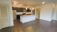 This single-floor space showcases a thoughtfully designed open-concept layout, anchored by a contemporary kitchen that serves as the heart of the living area. The kitchen features bold dark cabinetry with sleek hardware, contrasted by a light granite-topped central island equipped with an undermount sink — an ideal surface for both meal preparation and casual dining. Stainless steel appliances, including a refrigerator, microwave, and range, add a polished, modern touch. The kitchen is illuminated by a combination of track lighting along the ceiling and elegant pendant lights suspended above the island, creating layered, functional lighting throughout.

Rich, dark hardwood-style flooring extends seamlessly across the open space, lending warmth and continuity to the interior. A mosaic tile backsplash adds texture and visual interest behind the countertops. Visible through a doorway, a built-in shelving unit offers practical storage options. The open floor plan connects the kitchen to the broader living area, creating a spacious and versatile environment. The overall aesthetic is clean, modern, and functional, well-suited for everyday living.