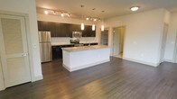 This single-floor space showcases a thoughtfully designed open-concept layout, anchored by a contemporary kitchen that serves as the heart of the living area. The kitchen features bold dark cabinetry with sleek hardware, contrasted by a light granite-topped central island equipped with an undermount sink — an ideal surface for both meal preparation and casual dining. Stainless steel appliances, including a refrigerator, microwave, and range, add a polished, modern touch. The kitchen is illuminated by a combination of track lighting along the ceiling and elegant pendant lights suspended above the island, creating layered, functional lighting throughout.

Rich, dark hardwood-style flooring extends seamlessly across the open space, lending warmth and continuity to the interior. A mosaic tile backsplash adds texture and visual interest behind the countertops. Visible through a doorway, a built-in shelving unit offers practical storage options. The open floor plan connects the kitchen to the broader living area, creating a spacious and versatile environment. The overall aesthetic is clean, modern, and functional, well-suited for everyday living.