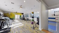 This comprehensive fitness center is designed to support a wide range of workout routines within a thoughtfully organized, single-floor layout. The space is immediately distinguished by its bold geometric mural wall art in shades of yellow, green, and teal, lending an energetic and modern aesthetic throughout the main workout areas.

The cardio zone features multiple treadmills and elliptical machines positioned near large windows that invite abundant natural light and views of the surrounding outdoor greenery. Ceiling fans overhead contribute to a comfortable workout environment. A dedicated alcove houses recumbent bikes alongside a wall-mounted flat-screen television, offering a more relaxed exercise setting.

The strength training area is well-stocked with a dumbbell rack, medicine ball tower, weight bench, and cable resistance machines, providing versatile options for both beginners and advanced users. A freestanding punching bag station in a separate open zone adds a functional space for cardio boxing and martial arts training.

Connecting to the fitness facility is a full indoor basketball court, notable for its high ceilings, bright overhead lighting, tiled flooring with regulation court markings in blue, and a bold blue accent stripe running along the block walls. French doors and window blinds throughout the fitness areas offer flexible light control and convenient outdoor access. The consistent grey-and-yellow patterned carpet flooring ties the various workout zones together cohesively.