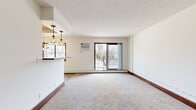 This single-floor unit offers a practical and efficient layout across a combined living, dining, and kitchen space, one bedroom, and a hallway. The open living and dining area is carpeted and bathed in natural light from large sliding glass doors that open to a balcony, providing outdoor views of the surrounding neighborhood. A wall-mounted air conditioning unit and baseboard heating ensure year-round comfort. The textured ceiling adds a classic touch throughout the main living spaces. The galley-style kitchen features warm wood cabinetry, stainless steel appliances including a full-size refrigerator and gas range, a drop sink, and durable light-toned vinyl plank flooring beneath a drop-panel ceiling with recessed lighting. The bedroom is a well-proportioned retreat with plush carpeting, a picture window framing outdoor tree views, and baseboard heating. A standout feature of the bedroom is the built-in dark wood cabinetry with open shelving, enclosed storage, and an integrated desk area, offering both organization and workspace. The hallway connects the rooms efficiently, featuring additional built-in cabinetry and countertop space. Consistent dark wood trim and baseboards throughout tie the space together with a cohesive, grounded aesthetic.