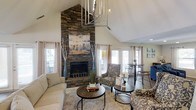 Located at 100 Eclipse Dr in Cary, NC, this thoughtfully designed single-floor space makes a lasting impression from the moment you enter. The main living area commands attention with its soaring vaulted ceilings that amplify the sense of openness and volume throughout the room. A floor-to-ceiling stacked stone fireplace serves as the dramatic focal point, adding a rustic yet refined character to the space. An artistic chandelier with an organic, sculptural design descends from the peak of the vaulted ceiling, contributing a distinctive decorative element that complements the natural textures found throughout. Large windows dressed with soft, neutral curtains line the walls, bathing the interior in generous natural light while offering views of the surrounding neighborhood. The living area is thoughtfully arranged with comfortable seating, round wooden coffee tables, and patterned accent chairs, creating a warm and welcoming atmosphere. Hardwood flooring anchors the space with warmth and durability. A bistro-style table and chairs in the background provide a casual seating nook, adding versatility to the layout. The overall ambiance is cozy yet airy, blending natural materials with curated décor to create an inviting and functional living environment.
