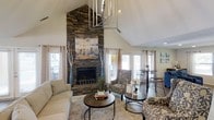 Located at 100 Eclipse Dr in Cary, NC, this thoughtfully designed single-floor space makes a lasting impression from the moment you enter. The main living area commands attention with its soaring vaulted ceilings that amplify the sense of openness and volume throughout the room. A floor-to-ceiling stacked stone fireplace serves as the dramatic focal point, adding a rustic yet refined character to the space. An artistic chandelier with an organic, sculptural design descends from the peak of the vaulted ceiling, contributing a distinctive decorative element that complements the natural textures found throughout. Large windows dressed with soft, neutral curtains line the walls, bathing the interior in generous natural light while offering views of the surrounding neighborhood. The living area is thoughtfully arranged with comfortable seating, round wooden coffee tables, and patterned accent chairs, creating a warm and welcoming atmosphere. Hardwood flooring anchors the space with warmth and durability. A bistro-style table and chairs in the background provide a casual seating nook, adding versatility to the layout. The overall ambiance is cozy yet airy, blending natural materials with curated décor to create an inviting and functional living environment.