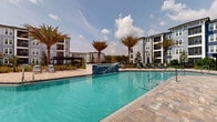 This expansive outdoor amenity area is designed to deliver a resort-like experience within a modern residential community. At its centerpiece is a generously sized swimming pool with vivid turquoise water, framed by a decorative tiled water feature and accessible via stainless steel entry rails. The pool deck is finished in warm-toned paver stone, providing ample space for sunbathing and outdoor lounging. A well-appointed sun deck lines one side of the pool and is furnished with cushioned chaise lounges, offering residents a comfortable place to relax in the open air. Adjacent to the pool is a covered pergola structure with a sleek dark metal frame, beneath which a central fire pit is surrounded by Adirondack-style chairs—ideal for evening gatherings. Tropical palm trees are thoughtfully integrated throughout the landscape, enhancing the resort ambiance and providing natural shade. A low-profile clubhouse building with large windows and bold blue and gray exterior accents anchors one end of the space, offering an architectural focal point and additional resident amenities. Manicured greenery, decorative lighting fixtures, and a fully fenced perimeter complete the polished, welcoming atmosphere of this outdoor community hub.
