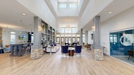 Located at Park Commons Drive in Knightdale, NC, this impressive single-floor community clubhouse offers a generously proportioned and thoughtfully designed amenity space. The grand entrance opens into a soaring two-story lobby distinguished by dramatic skylights, clerestory windows, and stacked-stone column bases that anchor the open floor plan. Light wood-style flooring flows seamlessly throughout, complementing a neutral palette of grays and whites accented by bold navy blue feature walls.

The central lounge area is furnished with plush velvet seating and elegant pendant lighting, creating an inviting gathering space bathed in natural light from the expansive window wall and glass entry doors. Adjacent, a cozy lounge nook features a floor-to-ceiling stacked-stone fireplace with a substantial slate mantel — a striking architectural focal point — accompanied by a wall-mounted TV and comfortable seating.

The games area includes a full-size billiards table and casual dining seating, while the kitchen bar showcases a granite waterfall-edge island with bar stools and pendant lighting overhead. A well-equipped fitness center occupies a dedicated wing. Curated artwork and decorative built-in shelving add refined character throughout this versatile, amenity-rich community space.