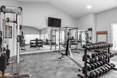 Located at 195 S Fifth W in Rexburg, Idaho, this fitness center offers a comprehensive workout facility within a single, open-plan space. The room features vaulted ceilings that lend a sense of openness and airiness, complemented by recessed lighting and neutral gray walls that create a clean, modern aesthetic. Durable carpet tile flooring covers the entire area, providing both comfort and traction underfoot.

The facility is thoughtfully organized into distinct workout zones. A strength training area is equipped with a full rack of dumbbells, a power rack, a cable crossover machine, and multiple adjustable benches, catering to a wide range of resistance training needs. A dedicated cardio section houses treadmills, ellipticals, and upright bikes, all positioned beneath a wall-mounted flat-screen television. Large wall mirrors amplify the sense of space and support proper form during exercise.

Natural light enters through multiple windows fitted with white blinds, as well as through a set of glass double doors that open to the outdoors, providing views of the surrounding greenery. Coat racks with benches near the entrance offer a convenient area to store personal items. The space is finished with clear emergency exit signage, ensuring a safe and organized environment for all users.
