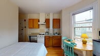 This single-floor studio in Cambridge, MA offers a well-organized open layout that efficiently combines living, sleeping, and cooking areas. The main room features warm hardwood floors and is lined with multiple double-hung windows on three sides, allowing abundant natural light and views of surrounding greenery. A curved bay-style wall with windows anchors one end of the room, lending architectural character to the space.

The kitchen area is fully equipped with warm-toned oak cabinetry, granite-style countertops, a tiled backsplash, a four-burner electric range with overhead ventilation, and a stainless steel sink. A compact refrigerator provides additional storage convenience. Adjacent to the kitchen, a small dining table with teal chairs sits neatly by a window, maximizing the use of available space.

The living and sleeping area includes a built-in wooden shelving unit that spans floor to ceiling, offering practical storage and display space. A cast-iron radiator provides heating, and a ceiling fan with light adds airflow and ambiance.

The bathroom features white tile walls, a vanity with storage, a toilet, and a full tub with a sliding glass shower enclosure, completing this functional and charming Cambridge studio.