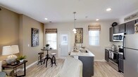 This single-floor 1-bedroom, 1-bathroom residence offers a well-designed open-concept layout that seamlessly connects the kitchen and dining areas, creating a functional and inviting living environment. The kitchen is a standout feature, showcasing a sleek island with a light granite countertop, an undermount sink, and pendant lighting that adds a polished, modern touch. Dark cabinetry provides a striking contrast against the lighter surfaces, contributing to a contemporary aesthetic. The dining area is efficiently arranged with a bistro-style high table and matching stools, positioned near a glass-paneled door that invites natural light into the space. Multiple windows dressed with patterned fabric valances and white blinds allow for ample daylight while maintaining privacy. Light wood-toned flooring runs throughout, enhancing the sense of openness and warmth. Neutral warm-beige walls serve as a versatile backdrop, complemented by tasteful wall art and decorative accents. Recessed ceiling lighting ensures the space remains bright and well-lit throughout the day. Overall, this residence combines practical design with modern finishes to deliver a comfortable and stylish single-floor living experience.