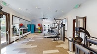 This fitness center is a comprehensively equipped, single-floor facility designed to support a full range of workout routines. The space features a modern, open layout with a patterned carpet flooring that defines distinct workout zones throughout the room.

The cardio area is lined with treadmills, elliptical trainers, stationary bikes, and stair climbers, each equipped with individual monitors for entertainment during workouts. Large windows along one wall allow ample natural light to fill the space and offer views of an outdoor pergola area, creating a bright and motivating environment.

The strength training section houses a variety of cable machines and multi-use weight stations, providing options for both upper and lower body conditioning. A vibrant yellow accent wall anchors this area, adorned with fitness reference charts, adding both visual energy and a functional resource for users.

A dedicated functional fitness area includes a colorful medicine ball rack, resistance accessories, and a large wall-mounted mirror that enhances the sense of space. An eye-catching landscape mural serves as a backdrop, adding an artistic element to the workout environment.

Ceiling fans throughout the facility ensure comfortable air circulation, and recessed lighting provides consistent, even illumination across the entire space. The overall design balances functionality with an inviting aesthetic, making it a well-rounded fitness destination.