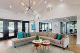 This thoughtfully designed single-story space blends contemporary style with functional amenities, creating an inviting environment suited for both relaxation and productivity. The heart of the space is a generously sized lounge area anchored by soaring vaulted, beamed ceilings that lend an airy, open feel. A dramatic sputnik-style chandelier serves as a focal point overhead, complementing the crisp white walls and warm herringbone-patterned hardwood flooring below. A curved, tufted sectional sofa in neutral tones, accented with teal and gray throw pillows, surrounds a pair of round wood-and-gold side tables, establishing a welcoming gathering atmosphere. A classic white fireplace with a tile surround and wall-mounted television anchors one wall, while black-framed French doors along the opposite wall flood the space with natural light and provide direct views and access to an outdoor area. Adjacent to the lounge, a refreshment station features white cabinetry with quartz countertops, a built-in beverage refrigerator, coffee station, microwave, water dispenser, and a full-size stainless steel refrigerator. Glass-walled private office spaces with black-framed partitions offer a sleek, professional setting visible from the main lounge, ensuring both connectivity and privacy throughout the space.