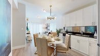 This thoughtfully designed single-floor residence offers a cohesive blend of modern aesthetics and functional living. The open-concept layout seamlessly connects the living room, dining area, and kitchen, creating a welcoming, airy atmosphere. The living room features large sliding glass doors dressed with sheer white curtains and horizontal blinds, allowing abundant natural light to fill the space. A wall-mounted television, a plush ivory sofa, a tufted ottoman, and rich leather accent chairs complete the inviting lounge area.

The dining space is anchored by an elegant round table surrounded by tufted velvet chairs with gold-toned legs, illuminated by a striking mid-century modern brass chandelier. The kitchen showcases crisp white cabinetry, dark countertops with a reflective backsplash, and stainless steel appliances.

Two bedrooms offer comfortable retreats, each with light wood-look flooring, generous natural light from well-dressed windows, built-in closets with louvered doors, and ample space for a desk or vanity area. The bathroom is finished with large-format tile flooring and a clean white vanity topped with a dark countertop.

A central hallway connects all rooms, finished in the same bright white palette that unifies the entire space, giving the home a polished, contemporary character throughout.