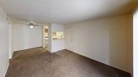 This single-floor residential unit offers a practical and functional layout designed for comfortable everyday living. The living area is generously sized, featuring wall-to-wall carpet, neutral cream-toned walls, a textured ceiling, and a ceiling fan. A pass-through window connects the living space to the kitchen, creating an open feel between the two areas. The front entry door opens directly into this main living space.

The kitchen is efficiently arranged in a galley-style configuration, with warm wood-toned cabinetry, light laminate countertops, vinyl flooring, and a full-size refrigerator. A pantry or utility closet is accessible from within the kitchen.

The primary bedroom is well-proportioned with plush carpet, a textured ceiling, and a sliding glass door fitted with vertical blinds that provides access to an outdoor space, allowing for natural light and ventilation. A second bedroom features a standard window with blinds, carpet flooring, and the same neutral color palette carried throughout the unit.

The bathroom includes a full-size soaking tub and shower combination with white tile surround, a toilet, a single-sink vanity with wood cabinetry, and durable tile flooring. Throughout the unit, the consistent use of neutral tones and carpet creates a cohesive, move-in ready atmosphere.