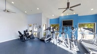 This fitness center is designed to provide a functional and energizing workout environment. The space features soaring vaulted ceilings that lend an open, airy feel, complemented by recessed lighting and a large ceiling fan that keeps the area comfortable during exercise. A bold blue accent wall serves as a striking focal point and adds a vibrant, motivating energy to the room. The durable, cushioned blue flooring provides a supportive surface underfoot, ideal for a high-traffic fitness environment. Large windows along the walls invite generous natural light and offer views of the surrounding greenery, creating a refreshing atmosphere. The cardio equipment selection is well-rounded, featuring treadmills, elliptical trainers, and stationary bikes arranged to allow for ample space between machines. A wall-mounted flat-screen television is positioned for easy viewing during workouts. The overall layout is thoughtfully organized, maximizing usable floor space while maintaining an uncluttered and welcoming environment. Whether used for a quick cardio session or a longer training routine, this fitness center offers a clean, modern, and well-lit space conducive to an effective workout experience.