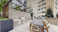 This expansive outdoor terrace is thoughtfully designed to offer a verdant urban retreat amid a striking city backdrop. A generous stretch of lush green lawn serves as the centerpiece, bordered by large, charcoal-toned rectangular planters that house mature trees, providing natural shade and a sense of enclosure. String lights are strung between poles above the lawn, lending a warm ambiance for evening gatherings.

A pergola-covered zone anchors one end of the terrace, housing a fully equipped outdoor kitchen with multiple stainless steel grills, ample counter space, and a long dining table with wicker and wood-frame chairs — ideal for group dining and entertaining. Concrete masonry walls define the perimeter, with raised planter beds along one edge filled with dense, manicured shrubs that add greenery and privacy.

The terrace connects directly to an interior space through floor-to-ceiling glass doors and windows, seamlessly blending indoor and outdoor environments. Surrounding high-rise buildings frame the space, reinforcing its urban character. The clean palette of light tile pavers, grey masonry, and modern metal detailing creates a cohesive, contemporary aesthetic throughout.