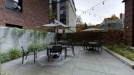 This outdoor patio courtyard offers a thoughtfully designed social gathering space nestled between urban architecture. The ground is finished with large-format concrete pavers, providing a clean and modern foundation throughout the space. Multiple dining sets, each accompanied by a large market umbrella, are arranged to offer comfortable and semi-private seating areas suited for both small and larger groups. Overhead, a canopy of globe string lights spans the courtyard, lending warmth and ambiance during evening hours. The perimeter is defined by a combination of rich brick walls, a horizontal wood privacy fence, and a decorative metal railing, which together create an enclosed yet open-air atmosphere. Climbing greenery and planted borders line the walls, softening the hardscape and introducing a natural element to the urban setting. Adjacent multi-story buildings with large industrial-style windows rise on either side, framing the courtyard and adding an architectural context characteristic of a contemporary mixed-use or residential development. The overall ambiance is inviting and functional, balancing privacy with openness, and making it a versatile outdoor amenity suitable for casual relaxation or social gatherings.
