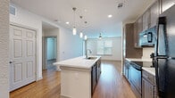 This single-floor residence showcases a thoughtfully designed open-concept layout that seamlessly connects the kitchen and living areas, creating a spacious and airy atmosphere. Warm hardwood-style flooring flows consistently throughout, anchoring the clean, contemporary aesthetic.

The kitchen is a standout feature, centered around a large island with a white countertop and an integrated sink, illuminated by a trio of sleek pendant lights. Dark-toned cabinetry with modern hardware lines the walls, complemented by black appliances including a refrigerator, electric range, built-in microwave, and dishwasher. Recessed lighting adds to the bright, functional ambiance.

The open living area extends naturally from the kitchen, benefiting from a ceiling fan and a window that invites natural light and outdoor views. The generous, unobstructed floor space offers excellent versatility for furnishing and arrangement.

The bedroom is a calm, well-proportioned room featuring large windows with blinds that frame views of a private balcony and surrounding greenery, flooding the space with natural light.

The bathroom is finished with a wide vanity topped with a light stone-style countertop, a drop-in sink, dark wood cabinetry below, and a walk-in glass-enclosed shower. A large mirror enhances the sense of space. Together, these elements create a cohesive, modern living environment with practical and aesthetic appeal.