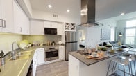 This single-floor residence combines contemporary design with practical functionality, offering a comfortable and visually appealing living environment. The kitchen serves as a standout feature, showcasing crisp white cabinetry, a built-in wine rack, and a full suite of stainless steel appliances, including a gas cooktop island, wall oven, microwave, and refrigerator. A bold stainless steel range hood anchors the space above the central island, which provides additional prep space and a neutral gray countertop surface that complements the overall palette. Light wood-style flooring, recessed lighting, and a green-toned tile backsplash add warmth and character to the kitchen, which flows seamlessly into the adjacent living and sleeping areas. The space is bathed in natural light, contributing to an open and airy atmosphere throughout. A private outdoor balcony extends the living space outward, offering expansive views of a lush desert landscape dotted with mature palm trees, manicured green spaces, and a well-maintained parking area below. In the distance, a mountain range frames the horizon beneath wide open skies, providing a dramatic and picturesque backdrop that enhances the overall appeal of this thoughtfully designed residence.