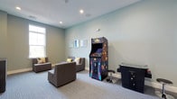 This single-floor amenity space is thoughtfully designed to offer both recreation and wellness in a clean, modern environment. The interiors are finished with soft sage-green walls, neutral carpeting, and recessed lighting throughout, creating a calm and inviting atmosphere.

The game lounge serves as a central gathering area, featuring comfortable modular gray sofas accented with colorful throw pillows and framed wall art. A standout upright arcade cabinet adds a retro-entertainment focal point to the room, complemented by an additional tabletop game unit nearby. A wall-mounted flat-screen television with a media console and speaker setup provides an additional entertainment option. Natural light filters in through large windows, brightening the space considerably.

Adjacent to the lounge, a corridor with light wood-framed glass doors leads into a well-equipped fitness center. The gym area features hardwood-style flooring, mirrored walls, and a range of exercise machines visible through the transparent doors, offering an open and motivating workout environment.

The overall design of this space balances functionality with comfort, making it well-suited for socializing, gaming, and maintaining an active lifestyle. The consistent color palette and contemporary finishes tie the various areas together into a cohesive and appealing amenity suite.