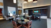 This sophisticated single-floor community lounge is designed to impress, combining contemporary aesthetics with a warm, inviting atmosphere. The space is anchored by dramatically high ceilings with exposed beam accents and recessed lighting that create an open, airy feel, while large pendant light fixtures add a refined finishing touch overhead.

The layout is thoughtfully divided into multiple functional zones. A casual seating area features a plush sofa and accent tables, ideal for relaxed conversation. Adjacent to it, several round dining tables paired with rich brown leather chairs provide comfortable settings for dining or social gatherings. Sleek dark tabletops and carpeted flooring in neutral tones tie the aesthetic together seamlessly.

Toward the back of the space, a bar area is framed by a striking accent wall of natural wood paneling, complemented by built-in shelving and bar-height seating. A mounted flat-screen television above the bar adds an entertainment focal point, while a billiards table nearby reinforces the lounge's recreational appeal.

The overall ambiance balances modern sophistication with a club-like warmth, achieved through a dark and neutral color palette, layered lighting, and carefully chosen furnishings. The space is well-suited for both casual socializing and organized gatherings.