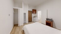 This studio unit is situated at 710 S Broadway in the heart of Downtown Los Angeles, offering a efficiently designed single-floor layout. The main living area features light wood laminate flooring, crisp white walls, and notable vertical space thanks to tall ceilings that lend an open, airy feel to the compact footprint. A window allows natural light into the sleeping area, which comfortably accommodates a full-size bed alongside a wooden nightstand. A wall-mounted heating unit provides climate control throughout the space.

The kitchenette is neatly tucked into one corner of the living area, equipped with overhead wood-finished cabinetry, a compact countertop range, and a mini refrigerator — providing essential cooking functionality within a streamlined design. Adjacent to the kitchenette, a reach-in closet with built-in shelving offers convenient storage solutions.

The private bathroom is clean and functional, featuring a pedestal sink with chrome fixtures, a wall-mounted mirror, and a corner stand-up shower enclosed with a glass and chrome sliding door. Light tile flooring ties the bathroom's neutral palette together.

The unit's straightforward, no-frills design makes it a practical option for urban living in one of Los Angeles's most accessible and well-connected neighborhoods.