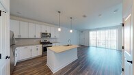 Located on Woodmoor Drive in Lombard, Illinois, this single-floor unit offers a thoughtfully designed open-concept layout that seamlessly connects the kitchen and living area. The kitchen is a standout feature, equipped with stainless steel appliances including a gas range, microwave, and refrigerator, all complemented by crisp white shaker cabinetry and granite countertops. A central island with an undermount sink serves as both a functional workspace and a gathering point, illuminated by pendant lighting overhead. Rich, dark-toned hardwood-style flooring runs throughout the main living areas, adding warmth and continuity to the space.

The living area is generously sized and bathed in natural light from a large sliding glass door that opens to a private balcony, offering outdoor views. The bedrooms feature soft carpeting and windows with blinds, providing a comfortable and quiet retreat. A well-appointed bathroom includes a granite-topped vanity with white cabinetry, a tiled tub-shower combination, and wood-style flooring. The hallway area provides ample closet storage, and the overall color palette of whites and neutrals contributes to a clean, modern, and inviting ambiance throughout.