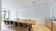 This thoughtfully designed single-floor amenity space blends modern elegance with functional versatility. The centerpiece of the space is a striking conference room anchored by a grand live-edge wood table with a luminous turquoise resin inlay, surrounded by leather and wood chairs. Light hardwood flooring, crisp white walls, and recessed ceiling lighting create a clean, focused atmosphere, while floor-to-ceiling windows at one end invite natural light and connect the room to the outdoors. A built-in credenza with warm wood cabinetry and a wall-mounted display screen complete the conference area, making it ideal for presentations and meetings. Adjacent to the conference room, the open lounge and café area offers a welcoming, resort-inspired environment. Structured around white columns, the lounge features woven armchairs, a café bar with a blue stone countertop and bar seating, and carefully placed indoor trees that add a natural, organic warmth to the interior. Patterned stone tile flooring in muted golds and grays defines the lounge zone, while recessed lighting and wall sconces contribute to a refined, inviting ambiance. Together, these spaces offer a seamless balance of professional functionality and relaxed sophistication.