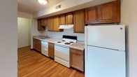 This single-floor residence presents a straightforward, functional layout with a clean and neutral aesthetic throughout. The living area is generously proportioned, featuring wall-to-wall beige carpeting and smooth white ceilings with recessed lighting. Vertical blinds on both a sliding glass door and a standard window allow for adjustable natural light while maintaining privacy. Floor-level air vents provide climate control, and the walls are finished in a soft, neutral tone that lends the space a bright and open feel.

Transitioning toward the back of the unit, the flooring shifts to hardwood, leading into a compact but well-equipped kitchen. Rich, warm-toned wood cabinetry lines the upper and lower walls, complemented by light granite-style countertops. White appliances—including a refrigerator, electric range, dishwasher, and range hood—are neatly integrated. The kitchen is efficiently arranged for easy workflow.

The bathroom is clean and well-appointed, featuring a white vanity with a granite-style countertop and single basin sink, a large mirror, a tiled tub-shower combination, and hardwood flooring. A multi-bulb overhead light fixture provides ample illumination. Throughout, the unit maintains a consistent, understated design suited to practical, everyday living.