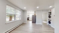 This single-floor unit offers a clean, modern interior characterized by light gray walls, light wood-tone luxury vinyl plank flooring, and recessed lighting throughout. The open living area is generously proportioned, receiving ample natural light through large windows fitted with blinds, and is equipped with baseboard heating. The space flows seamlessly toward the kitchen, which is separated by a partial wall partition. The kitchen is well-appointed with white shaker-style cabinetry, white subway tile backsplash, white countertops, and a full suite of stainless steel appliances including a gas range with hood, refrigerator, and dishwasher. A window above the sink brings in additional natural light. A hallway connects the living area to the private bedroom, which features dual windows on adjacent walls, filling the room with bright, diffused light, along with baseboard heating and continued vinyl plank flooring. The bathroom includes a white vanity with storage, a toilet, and dark tile flooring, with a window providing ventilation and light. A dedicated laundry closet houses a stacked washer and dryer unit, adding significant everyday convenience. The overall aesthetic is fresh, neutral, and move-in ready.