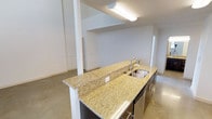This two-floor space offers a clean, modern aesthetic defined by an open layout and minimalist design. The main level showcases polished concrete floors that extend throughout, lending an industrial yet refined character to the space. At the heart of the main floor is a kitchen island topped with striking gold-veined granite countertops, complete with a double stainless steel sink and integrated dishwasher. Dark cabinetry beneath the island provides a bold contrast to the light stone surface above. The open-plan layout allows the kitchen to flow naturally into the surrounding living areas, which benefit from bright white walls and ample overhead lighting. A structural column near the island adds an architectural element while maintaining the sense of openness. The second floor is accessible from the main level, with the upper space visible from below, reinforcing the vertical scale of the interior. Clean sightlines, neutral tones, and durable finishes throughout create an environment that is both functional and visually cohesive. The understated design palette allows the architectural features and material choices to take center stage, making this a versatile and well-appointed space.