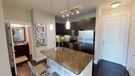 The kitchen is a standout feature of this single-floor space, combining functionality with a polished aesthetic. Dark espresso-toned cabinetry lines the walls, providing ample storage while contributing to a rich, contemporary look. A green tile backsplash adds a subtle pop of color, complementing the warm tones of the granite countertops that extend across the workspace and wrap around a central breakfast bar peninsula. The peninsula, finished with a white base and dark granite surface, offers casual seating and serves as a natural dividing point between the kitchen and the adjacent living area. Stainless steel appliances — including a French door refrigerator with an ice and water dispenser, an electric range, and an over-the-range microwave — provide a sleek, modern finish. Lighting is thoughtfully layered, with a track lighting system over the main kitchen area and elegant pendant lights hanging above the peninsula, ensuring the workspace is well-illuminated. Dark hardwood flooring runs throughout, tying the space together cohesively. Decorative wall mirrors near the entryway add depth and a stylish touch to the overall interior.