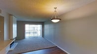This single-floor residence offers a clean and functional layout designed for comfortable everyday living. The main living area is immediately welcoming, characterized by a large sliding glass door framed by vertical blinds that fills the room with generous natural light while providing access to an outdoor balcony or patio. The space features a combination of plush wall-to-wall carpeting and rich hardwood flooring, creating a clear visual distinction between the living and transitional areas. A decorative pendant light fixture with a classic bowl shade and brass-toned hardware serves as a tasteful focal point, casting warm ambient light throughout the room. The walls are finished in a neutral tone, offering a versatile backdrop suited to a variety of interior styles. A textured popcorn ceiling adds a traditional touch characteristic of the home's construction era. White baseboards provide a crisp contrast along the perimeter of the room. The overall ambiance is bright, understated, and move-in ready, making it a practical and inviting space for a range of living arrangements.