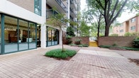 This single-floor space is situated at the base of a multi-story brick building, offering a seamless connection between interior and exterior environments. Large floor-to-ceiling glass doors and windows provide excellent visibility and allow natural light to flood the interior, creating an open and inviting atmosphere. The space opens directly onto a charming paved courtyard, featuring a classic brick-patterned walkway and carefully maintained landscaping. Young trees planted in structured ground beds, low shrubs, and neatly edged greenery contribute to a serene, park-like setting. A low brick perimeter wall defines the outdoor area, with a set of distinctive yellow steps leading up to a secondary pathway, adding a touch of character to the surroundings. Mature trees nearby provide natural shade and privacy, enhancing the overall ambiance. The building facade features dark-framed balconies on upper floors, reinforcing the architectural continuity of the property. This ground-level space is well-suited for commercial, retail, or communal use, benefiting from strong visual exposure, an abundance of natural light through its glass frontage, and direct access to the appealing outdoor courtyard environment.