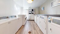 Located at 41 Dundonald Street in Toronto's Church-Wellesley Village, this shared laundry facility is a well-organized, single-floor amenity space designed for efficient everyday use. The room is laid out with rows of top-loading washing machines and front-loading dryers lining both sides, providing ample laundry capacity for building residents. A dedicated utility sink with a gooseneck faucet and white cabinetry offers additional functionality for hand-washing or pre-treatment tasks. A dark granite-style countertop runs along one wall, providing a convenient folding and sorting surface. The flooring throughout is warm-toned wood-look vinyl plank, lending a clean and modern aesthetic to the utilitarian space. Overhead flush-mount lighting ensures the room is bright and well-lit. A bulletin board near the window displays notices and building information, keeping residents informed. The space also features direct access to a shared washroom, clearly marked with signage, as well as a wall-mounted fire extinguisher and first aid kit for safety. A window at one end allows for natural light to enter. The overall design is functional, tidy, and well-maintained, reflecting a practical shared amenity space suited to a residential building.