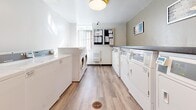Located at 41 Dundonald Street in Toronto's Church-Wellesley Village, this shared laundry facility is a well-organized, single-floor amenity space designed for efficient everyday use. The room is laid out with rows of top-loading washing machines and front-loading dryers lining both sides, providing ample laundry capacity for building residents. A dedicated utility sink with a gooseneck faucet and white cabinetry offers additional functionality for hand-washing or pre-treatment tasks. A dark granite-style countertop runs along one wall, providing a convenient folding and sorting surface. The flooring throughout is warm-toned wood-look vinyl plank, lending a clean and modern aesthetic to the utilitarian space. Overhead flush-mount lighting ensures the room is bright and well-lit. A bulletin board near the window displays notices and building information, keeping residents informed. The space also features direct access to a shared washroom, clearly marked with signage, as well as a wall-mounted fire extinguisher and first aid kit for safety. A window at one end allows for natural light to enter. The overall design is functional, tidy, and well-maintained, reflecting a practical shared amenity space suited to a residential building.