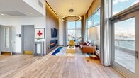 Located in East Boston, this sky-level amenity space delivers an exceptional combination of social, recreational, and wellness facilities, all framed by sweeping waterfront views of Boston Harbor and the city skyline. The open lounge and dining area features wide-plank hardwood flooring, floor-to-ceiling glass walls, and elegant nautical-inspired pendant lighting that hang above a rustic wood dining table surrounded by stylish seating. A striking herringbone-tiled accent wall anchors the living area, complete with a sleek linear fireplace beneath a mounted flat-screen TV, creating a warm and inviting focal point. The modern kitchen is outfitted with white cabinetry, a stainless steel refrigerator, built-in wall oven, and a dedicated workspace, providing full functionality for entertaining. The fitness center is well-appointed with professional-grade cardio equipment including treadmills, ellipticals, and cycling bikes, along with free weights and mirrored walls — all bathed in natural light from large windows overlooking the water. The outdoor deck extends the living space with large-format tile, railing seating, and direct panoramic views of the harbor and the Tobin Bridge, offering an unmatched outdoor retreat above the Boston skyline.