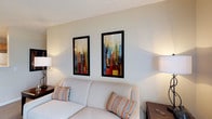 Located at 130 Cumberland Way in Pataskala, Ohio, this single-floor space offers a clean and welcoming environment designed for comfort and functionality. The living area sets a relaxed tone with a light-colored upholstered sofa accented by patterned throw pillows in warm earth tones, creating an inviting place to unwind. The walls are finished in soft, neutral hues that complement the overall calm ambiance of the space. A standout feature of the living area is the pair of bold, framed contemporary artworks displayed prominently on the wall — their vivid cityscape compositions in red, blue, green, and gold add a dynamic visual focal point that brings energy and color to the otherwise understated palette. Ambient lighting from table lamps on either side of the sofa contributes to a warm and comfortable atmosphere. The textured ceiling adds a subtle architectural detail that enhances the room's character. Overall, this space strikes a thoughtful balance between simplicity and style, making it a comfortable and visually engaging environment well-suited for everyday living in the Pataskala, Ohio community.
