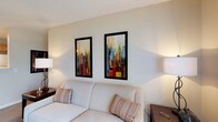 Located at 130 Cumberland Way in Pataskala, Ohio, this single-floor space offers a clean and welcoming environment designed for comfort and functionality. The living area sets a relaxed tone with a light-colored upholstered sofa accented by patterned throw pillows in warm earth tones, creating an inviting place to unwind. The walls are finished in soft, neutral hues that complement the overall calm ambiance of the space. A standout feature of the living area is the pair of bold, framed contemporary artworks displayed prominently on the wall — their vivid cityscape compositions in red, blue, green, and gold add a dynamic visual focal point that brings energy and color to the otherwise understated palette. Ambient lighting from table lamps on either side of the sofa contributes to a warm and comfortable atmosphere. The textured ceiling adds a subtle architectural detail that enhances the room's character. Overall, this space strikes a thoughtful balance between simplicity and style, making it a comfortable and visually engaging environment well-suited for everyday living in the Pataskala, Ohio community.
