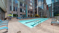 Located at 345 E Wacker Drive in the heart of Chicago, this outdoor pool deck offers a well-appointed recreational retreat set against a dramatic urban backdrop. The centerpiece of the space is a generously sized lap pool with clearly marked swim lanes, stainless steel entry rails, and crystal-clear water, designed to accommodate both serious swimmers and leisure use. Surrounding the pool, an expansive tiled deck provides ample space for relaxation, lined with rows of blue lounge chairs and accented by vibrant green and blue market umbrellas for shade. The deck is framed by the striking glass-and-steel facades of neighboring high-rise towers, creating a distinctly metropolitan atmosphere. A separate lounge area is tucked behind decorative railings, featuring additional seating, a grill station, and lush landscaped planters filled with flowering plants, ornamental grasses, and small trees, adding a touch of greenery to the urban setting. Colorful flower beds along the building's perimeter further enhance the outdoor ambiance. Large floor-to-ceiling glass doors connect the deck to the interior, seamlessly blending indoor and outdoor spaces. The overall design balances functionality with comfort, making this pool deck an exceptional urban amenity in one of Chicago's most dynamic neighborhoods.