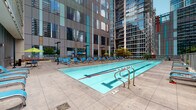 Located at 345 E Wacker Drive in the heart of Chicago, this outdoor pool deck offers a well-appointed recreational retreat set against a dramatic urban backdrop. The centerpiece of the space is a generously sized lap pool with clearly marked swim lanes, stainless steel entry rails, and crystal-clear water, designed to accommodate both serious swimmers and leisure use. Surrounding the pool, an expansive tiled deck provides ample space for relaxation, lined with rows of blue lounge chairs and accented by vibrant green and blue market umbrellas for shade. The deck is framed by the striking glass-and-steel facades of neighboring high-rise towers, creating a distinctly metropolitan atmosphere. A separate lounge area is tucked behind decorative railings, featuring additional seating, a grill station, and lush landscaped planters filled with flowering plants, ornamental grasses, and small trees, adding a touch of greenery to the urban setting. Colorful flower beds along the building's perimeter further enhance the outdoor ambiance. Large floor-to-ceiling glass doors connect the deck to the interior, seamlessly blending indoor and outdoor spaces. The overall design balances functionality with comfort, making this pool deck an exceptional urban amenity in one of Chicago's most dynamic neighborhoods.